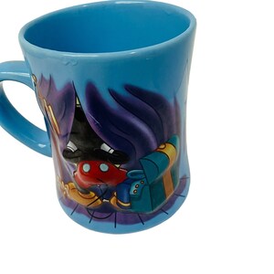 May include: A light blue ceramic mug with a cartoon character design. The mug has a curved shape and a handle. The design includes a character in a black outfit with red shorts and yellow shoes. The mug is approximately 10 cm tall.