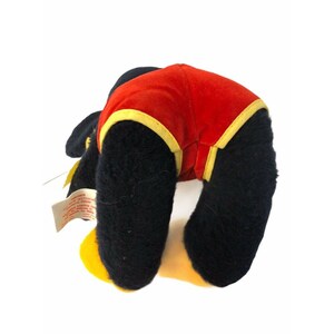 May include: A plush toy featuring a black body, red shorts with yellow trim, and yellow feet. The toy is bent over, with its rear facing the viewer. A tag is attached to the toy.