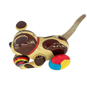 May include: Vintage wind-up toy dog with a brown and tan spotted body, red collar, and a metal tail. The toy has two multi-colored ball wheels. A classic collectible toy.