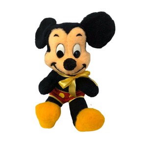 May include: A vintage Mickey Mouse plush toy. The plush features black fur, a tan face, large black ears, and yellow feet. Mickey wears red shorts with yellow buttons and a yellow bow tie. The toy is a classic Disney collectible.