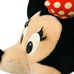 May include: Close-up of a plush Minnie Mouse head. The character has a light pink face, large white eyes with black pupils, and long black eyelashes. A red bow with white polka dots sits atop the head. The plush is black and cream-colored.