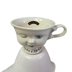 May include: A white ceramic cup with a face design, featuring a winking eye and a blue eye. The cup has a handle and the word "BAILEYS" is printed on the inside. The cup is placed on top of a white ceramic object.