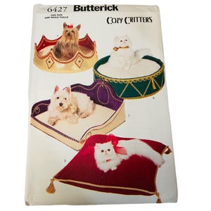 Puede incluir: Un patrón de costura Butterick para camas para mascotas Cozy Critters. El patrón presenta cuatro diseños de cama diferentes, incluyendo una cama en forma de corona, una cama redonda con ribete verde, una cama morada y beige y una cama de almohada roja.