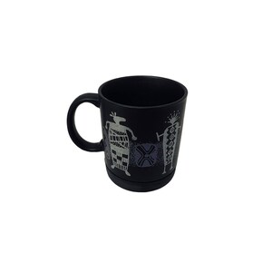May include: Black ceramic mug featuring stylized figures in white and gray. The mug has a handle and a decorative band at the base. The design includes geometric and human-like shapes, suitable for coffee or tea.