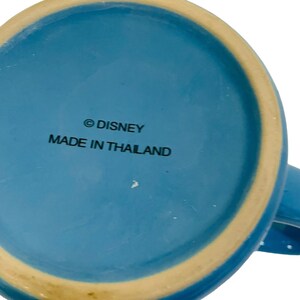 May include: A light blue ceramic mug with a cream-coloured rim. The base of the mug features the text "&copy; DISNEY MADE IN THAILAND" in black lettering. The mug is set against a white background.
