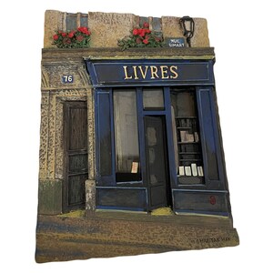 May include: A detailed miniature of a Parisian bookstore with a blue facade and the word "LIVRES" above the entrance. The model includes a door, window, and a sign that reads "RUE SIMART". Red flowers adorn the top of the building.