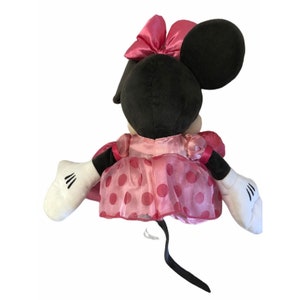 Pu&ograve; includere: Una bambola di peluche di Minnie Mouse, vista da dietro, con orecchie nere e un fiocco rosa. Indossa un vestito rosa a pois rossi e guanti bianchi con dettagli neri. Una coda nera si estende dalla bambola.