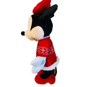 May include: A plush Minnie Mouse doll wearing a red sweater with white snowflake patterns, a matching red bow, and red shoes. The doll has black ears and a white fur trim on the sweater and skirt.