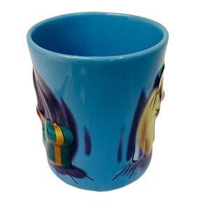 May include: A light blue ceramic mug with a cartoon design. The mug features a yellow and white character with black whiskers and a gift box. The mug has a smooth, glossy finish and a rounded shape.