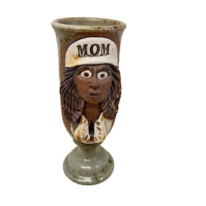 May include: A handmade ceramic goblet with a sculpted face of a woman wearing a hat with the word "MOM" on it. The goblet is brown, tan, and green, with a pedestal base. The face has large eyes and detailed hair.