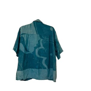 May include: Teal short-sleeved button-up shirt with a light teal abstract pattern. The shirt has a collared neckline and a relaxed fit. The pattern features organic shapes and a silhouette design. The shirt is made of a lightweight fabric.