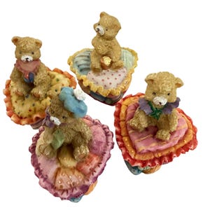 May include: Four small, decorative teddy bear figurines. Each bear is light brown and sits atop a heart-shaped cushion with ruffled edges. The cushions are various colors, including pink, yellow, and blue.