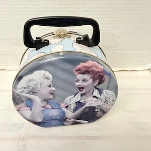 May include: Vintage metal lunchbox featuring a black handle and a clasp closure. The lunchbox has a light blue background with pink hearts. The front of the lunchbox displays a black and white image of two women.