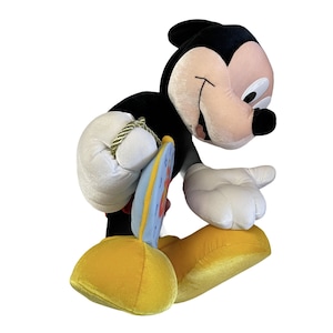 Puede incluir: Un peluche de Mickey Mouse en una pose juguetona. El juguete presenta un cuerpo negro, guantes blancos y zapatos amarillos. Sostiene un peque&ntilde;o objeto circular y colorido con un borde azul y detalles rojos. La cara del juguete es de color rosa claro.