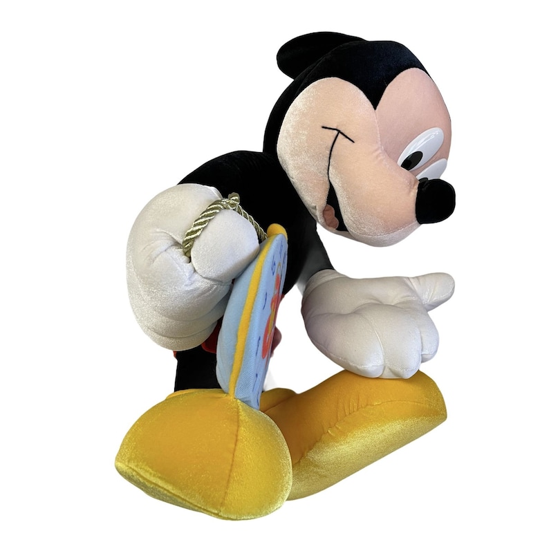 Puede incluir: Un peluche de Mickey Mouse en una pose juguetona. El juguete presenta un cuerpo negro, guantes blancos y zapatos amarillos. Sostiene un peque&ntilde;o objeto circular y colorido con un borde azul y detalles rojos. La cara del juguete es de color rosa claro.