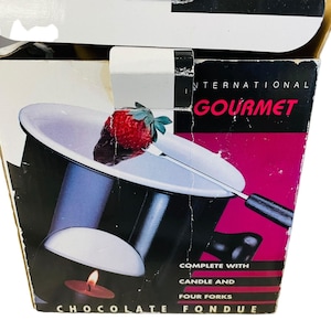 May include: A black and silver chocolate fondue set with a strawberry on a fork. The box reads "INTERNATIONAL GOURMET CHOCOLATE FONDUE" and includes a candle and four forks.
