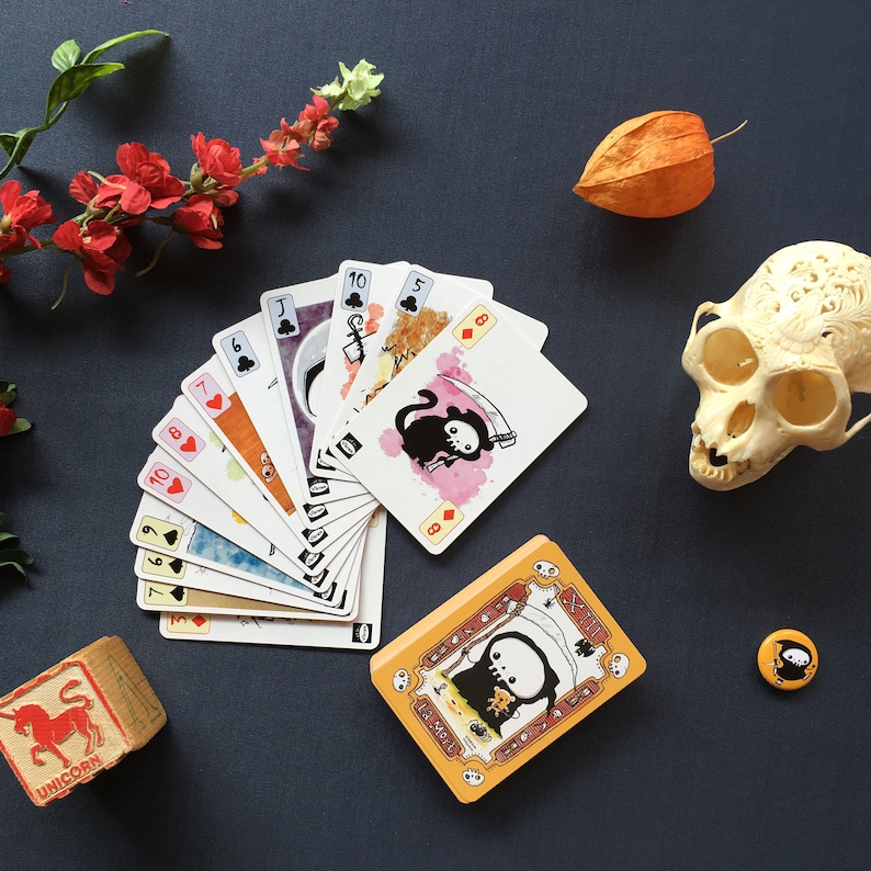 Puede incluir: Un juego de cartas con gatos negros y un dise&ntilde;o de calavera. Las cartas est&aacute;n dispuestas en forma de abanico sobre una superficie azul. Las cartas est&aacute;n numeradas del 6 al 10 e incluyen un Jack, una Reina y un Rey. Las cartas forman parte de un juego de cartas m&aacute;s grande con tem&aacute;tica de Halloween.