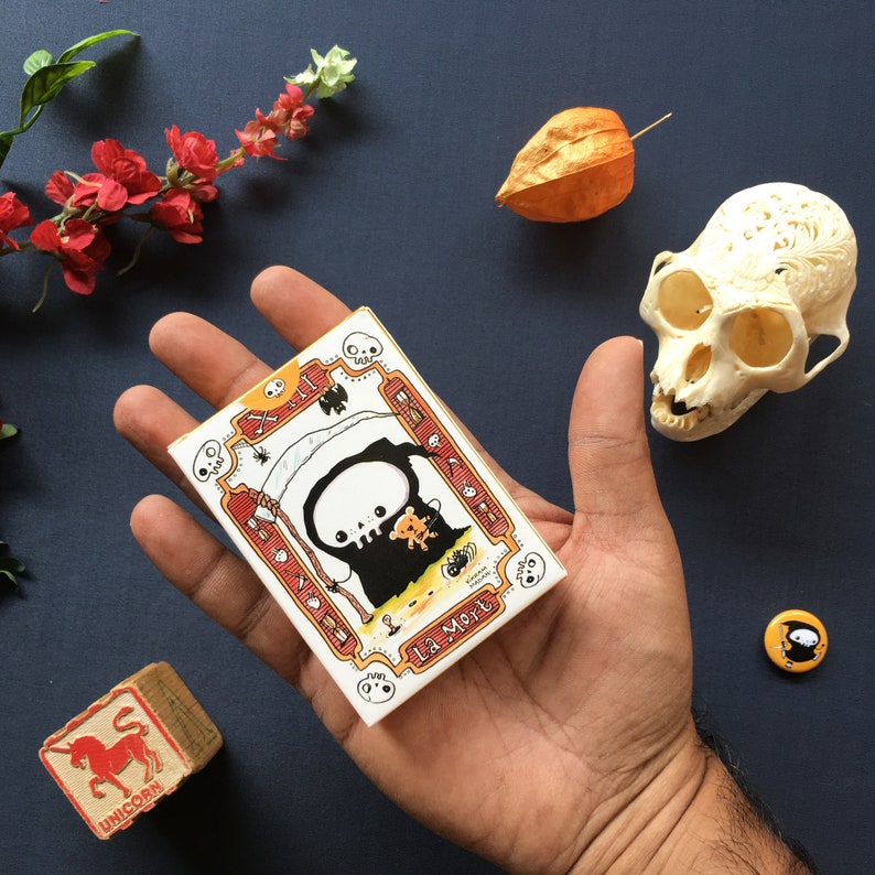 Puede incluir: Una tarjeta blanca con una ilustraci&oacute;n en blanco y negro de un esqueleto con una t&uacute;nica con capucha y sosteniendo un peque&ntilde;o oso. La tarjeta tiene acentos rojos y naranjas y el texto "La Mort" en negro.