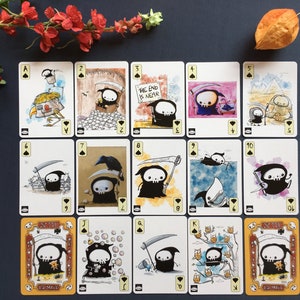 Puede incluir: Una baraja de cartas de juego con una caricatura de la Muerte en varias situaciones humor&iacute;sticas. Las cartas son del tama&ntilde;o est&aacute;ndar de las cartas de juego y presentan los palos est&aacute;ndar de picas, corazones, diamantes y tr&eacute;boles. Las cartas est&aacute;n impresas en cartulina blanca con tinta negra.