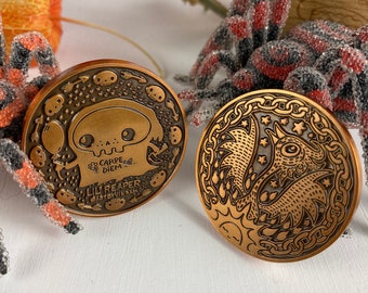 Puede incluir: Dos pines de metal de color cobre. El primer pin presenta una calavera de dibujos animados con el texto "Carpe Diem" y "Lil Reaper" debajo. El segundo pin presenta una ilustración detallada de una criatura mítica con estrellas y un sol.