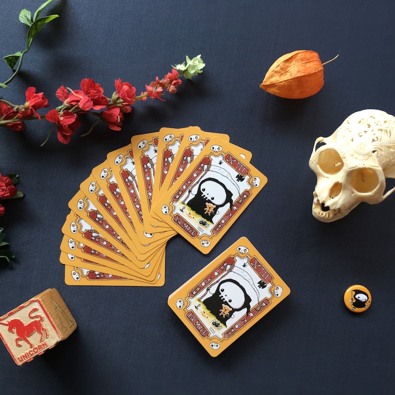 Puede incluir: Un juego de cartas amarillas con ilustraciones negras de un personaje esqueleto. Las cartas presentan un dise&ntilde;o de calavera y huesos cruzados y el texto "The Skull".