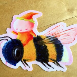 Könnte beinhalten: Ein Aufkleber mit einer Cartoon-Biene, auf der ein kleiner, rosa-häutiger Mensch mit einem roten Hut und einer gelben Krone reitet. Die Biene hat schwarz-gelbe Streifen, blaue Flügel und einen flauschigen Körper.