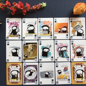 Puede incluir: Una baraja de cartas de juego con una ilustraci&oacute;n en blanco y negro de un esqueleto sosteniendo una guada&ntilde;a. Las cartas est&aacute;n numeradas del 1 al 13 e incluyen el As, el J, la Q y el K. Las cartas est&aacute;n todas en el mismo estilo, con un fondo negro y contornos blancos.