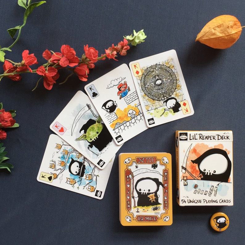 Puede incluir: Una baraja de cartas con una ilustraci&oacute;n en blanco y negro de un segador en la caja. La caja es amarilla y naranja con el texto "Lil' Reaper Deck" y "54 cartas de juego &uacute;nicas". Hay cinco cartas extendidas sobre una superficie azul, cada una con una ilustraci&oacute;n diferente.