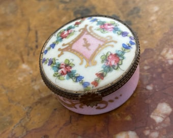 May include: A small, round, pink porcelain trinket box with a gold-trimmed lid. The lid is decorated with a white background and a pink diamond shape with gold trim. The diamond shape is surrounded by a floral pattern in shades of blue, green, and pink.