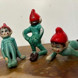 May include: Three ceramic figurines of elves with red hats and green bodies. The elves are in various poses, one is sitting, one is standing, and one is crawling.