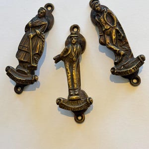 May include: Three brass door knockers featuring relief figures. The left depicts a person labeled MRS BARDELL, the center shows a figure labeled PERKER THE LAWYER, and the right displays a figure labeled OLIVER TWIST. Each has mounting holes.
