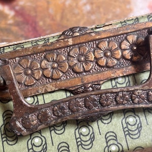 May include: A bronze-toned metal drawer pull featuring a rectangular backplate embossed with a repeating five-petal flower pattern and a matching hinged drop handle. The hardware displays a weathered finish with visible signs of age and wear.