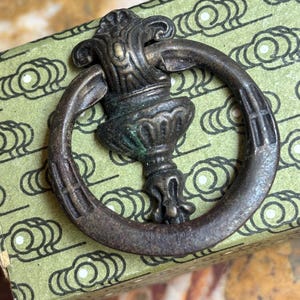 May include: A dark bronze-toned metal drawer pull featuring a circular ring attached to a decorative, ornate mounting plate with vertical fluting and a weathered finish, showing subtle hints of green oxidation on the central metal structure.