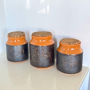 May include: Three ceramic kitchen storage jars with cork lids. The jars are two-tone, with a black base and orange top. The words "Salt", "Flour", and "Rice" are written on the front of each jar in a cursive font.