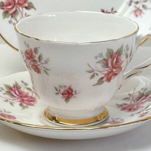 May include: A white porcelain teacup and saucer set featuring a delicate pink rose floral pattern with green leaves and small blue accents. The teacup has a scalloped rim and a gold-trimmed base, while the saucer edge is also finished in gold.