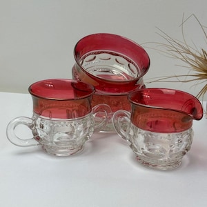 May include: A collection of clear glass serving items with ruby red rims. The set includes two handled cups and a creamer. The glass has a textured pattern. The items are arranged on a white surface.