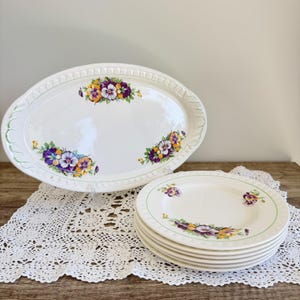 May include: An oval serving platter and a stack of five round plates, all made of white ceramic with a decorative embossed rim. Each piece features a colourful floral pattern of purple, yellow, and white pansies arranged in clusters on the surface.