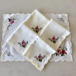 May include: A set of off-white fabric napkins and a matching rectangular placemat, each featuring scalloped edges and embroidered red rose motifs with green leaves and small blue flowers. The napkins are folded and stacked on the placemat.