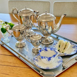 May include: A silver tea service set on a rectangular silver tray, including two teapots, a sugar bowl, and a creamer. A white porcelain teacup and saucer with blue floral patterns sit on the tray next to a small plate with cucumber sandwiches.