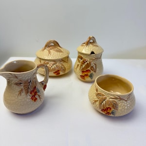 May include: A vintage ceramic cream and sugar set comprising a jug, two lidded pots, and a small bowl. The set has a textured beige finish with brown and red leaf and berry patterns. The jug has a handle and spout.