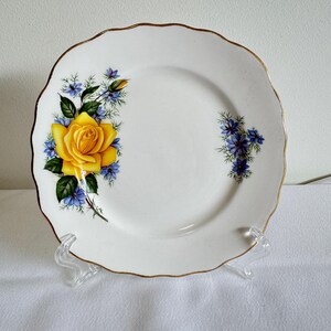 May include: White ceramic plate with a gold scalloped edge, decorated with a yellow rose and blue floral pattern. The plate is supported by a clear stand. A vintage-style plate, ideal for serving or display, with a diameter of approximately 20 cm.