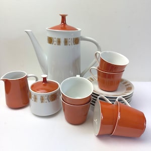 May include: A vintage white and orange ceramic coffee set. The set includes a tall coffee pot, cups, saucers, a creamer, and a sugar bowl. The coffee pot and sugar bowl have a decorative geometric pattern.