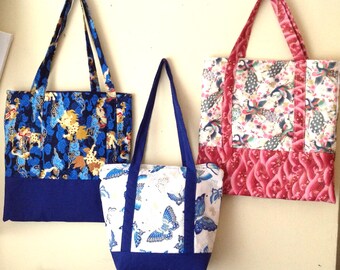 Könnte beinhalten: Drei Tragetaschen mit verschiedenen Stoffmustern. Die größte Tasche ist blau mit einem goldenen und weißen Muster, die mittlere Tasche ist weiß mit einem goldenen und blauen Schmetterlingsmuster und die kleinste Tasche ist pink mit einem weißen und blauen Pfauenmuster.