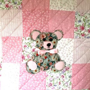 Op de afbeelding: Een patchwork quilt met een bloemen teddybeer applicatie. De quilt heeft vierkanten van roze, witte en bloemenstoffen. De teddybeer is gemaakt van bloemenstof en heeft een roze strik.