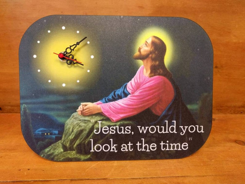 A rectangular novelty clock with a picture of Jesus praying to the heavens.  The clock face is to one side.  Jesus is not looking at it.  Text on the clock face says Jesus would you look at the time.