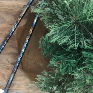 May include: Two twisted metal bar tools with a blue, purple, and brown iridescent finish. The tools are resting in a wooden bowl with green pine branches in the background.