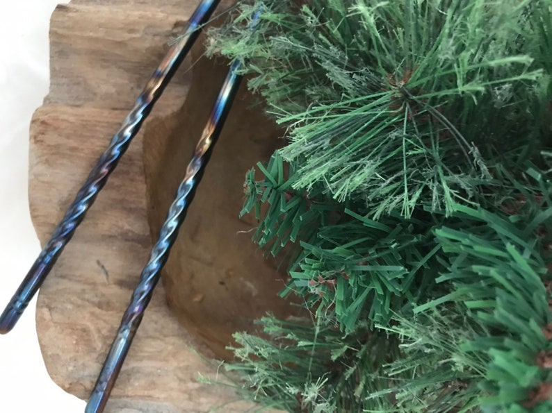May include: Two twisted metal bar tools with a blue, purple, and brown iridescent finish. The tools are resting in a wooden bowl with green pine branches in the background.