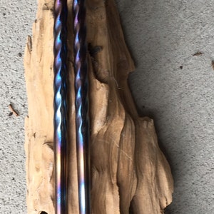 May include: A pair of iridescent blue and purple chopsticks with a twisted design. The chopsticks are resting on a piece of driftwood.
