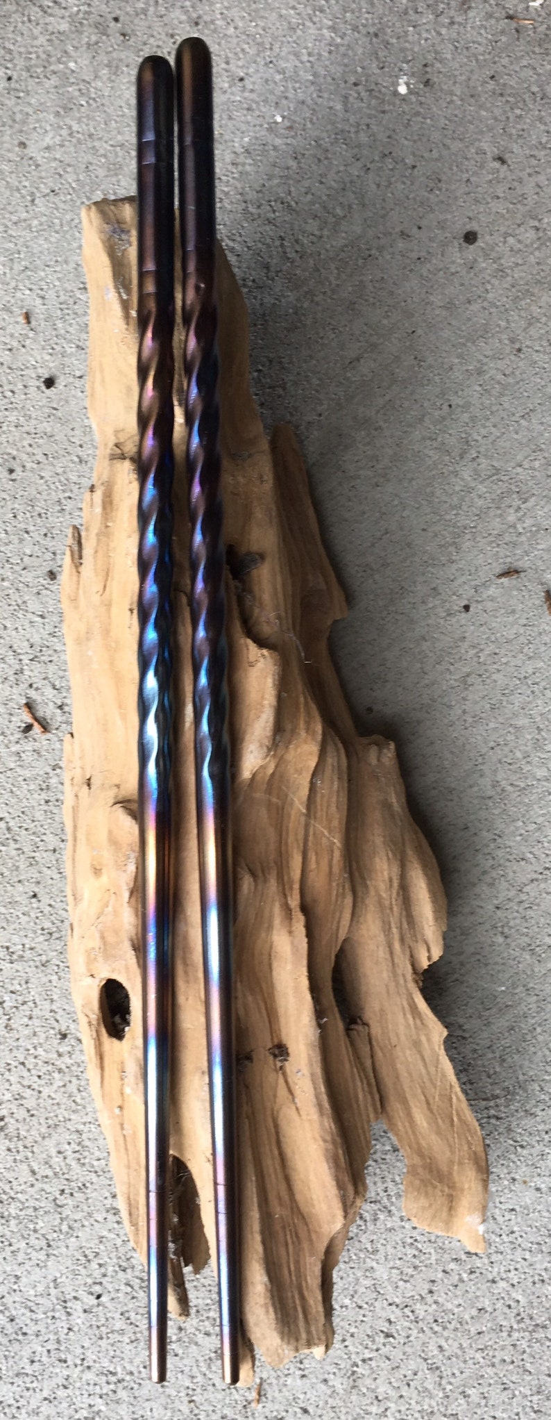 May include: A pair of iridescent blue and purple chopsticks with a twisted design. The chopsticks are resting on a piece of driftwood.