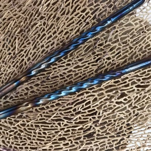 May include: Two blue and silver twisted metal chopsticks on a brown textured surface.